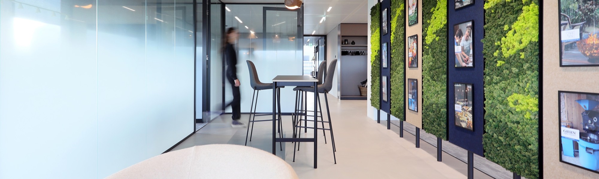 Moderne kantoorruimte met hoge tafel en krukken, glazen wanden, een groene plantenwand met fotolijsten en een persoon op de achtergrond.