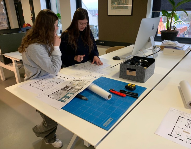 Twee vrouwen zitten samen aan een bureau in een kantoor en bekijken bouwtekeningen die op tafel liggen. Op het bureau liggen een snijmat, opgerolde tekeningen, stiften en een opbergbox met kantoorbenodigdheden. Eén van hen wijst naar de tekening terwijl z