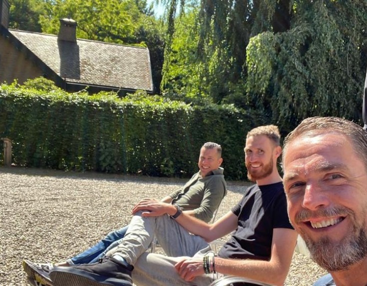 Drie mannen zitten ontspannen naast elkaar op een grindterras in de zon, met op de achtergrond een groene haag, hoge bomen en een landelijk huis met rieten dak. De man op de voorgrond maakt een selfie en glimlacht breed in de camera, terwijl de andere twe