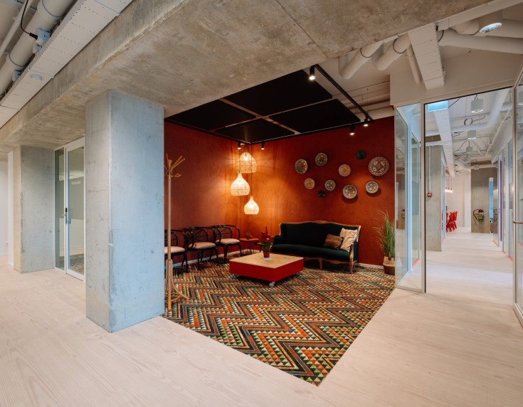 Moderne kantoorruimte met een gezellige zithoek in een nis met rode muur, bank en fauteuils op een kleurrijk patroonkleed, omringd door een lichte open werkruimte met glazen wanden.