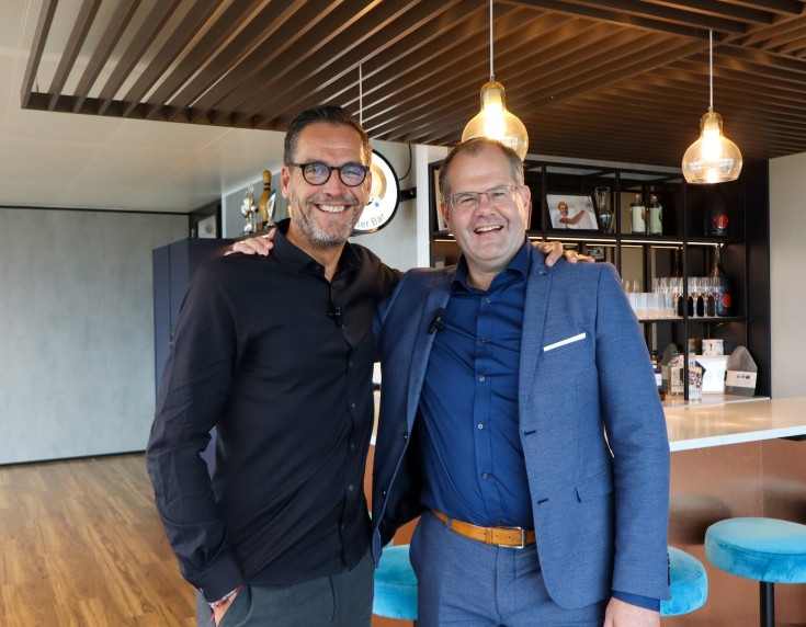 Twee mannen staan lachend naast elkaar in moderne kantooromgeving met bar en hanglampen.