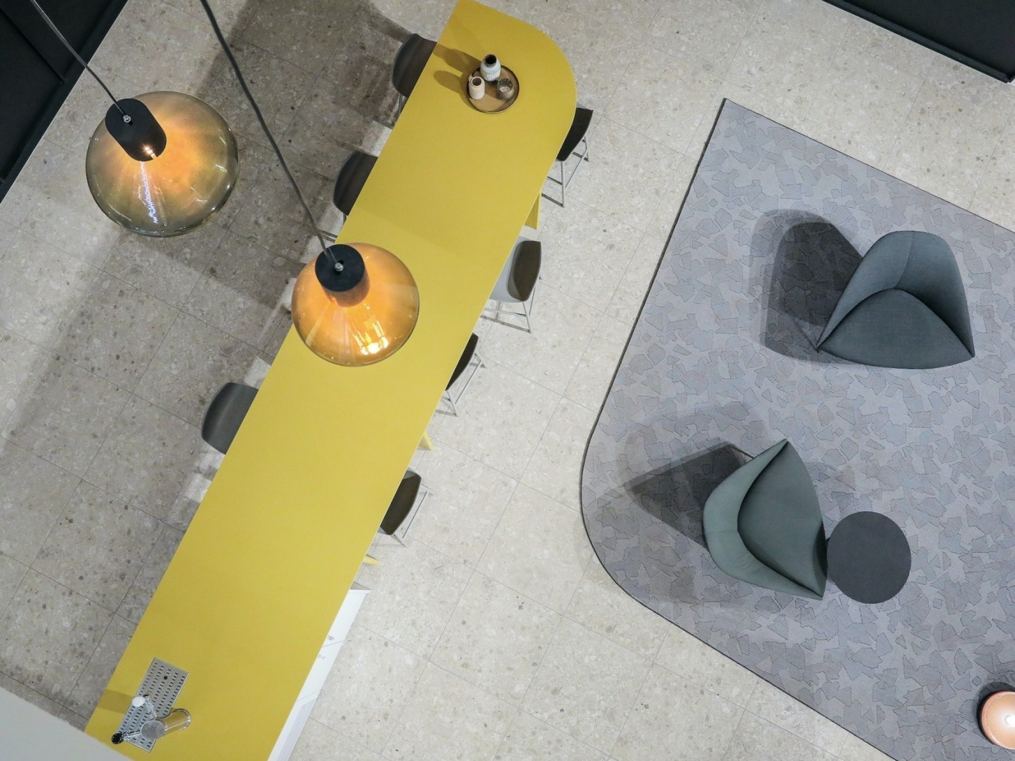 Bovenaanzicht van een moderne kantoorruimte met een lange gele tafel, hanglampen, stoelen en een zithoek met twee fauteuils op een grijs vloerkleed.