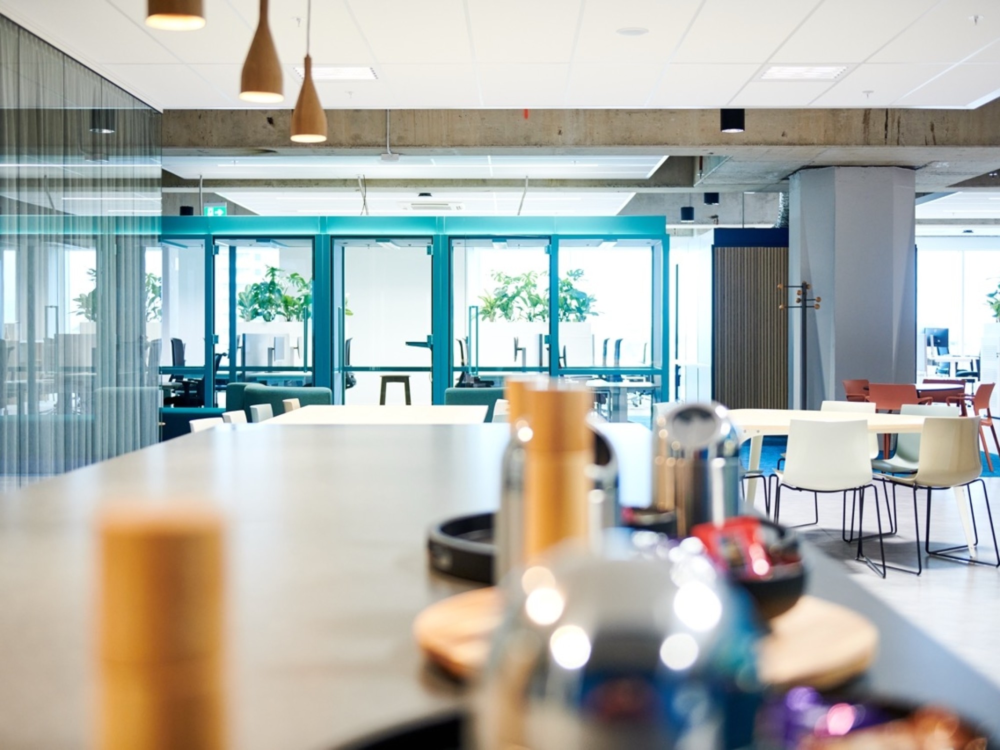 Een moderne kantoorruimte met een lange tafel op de voorgrond waarop dienbladen en bekers staan. Op de achtergrond zijn glazen deuren met turquoise kozijnen zichtbaar en verschillende tafels en stoelen in een open ontmoetingsruimte. De ruimte is licht en