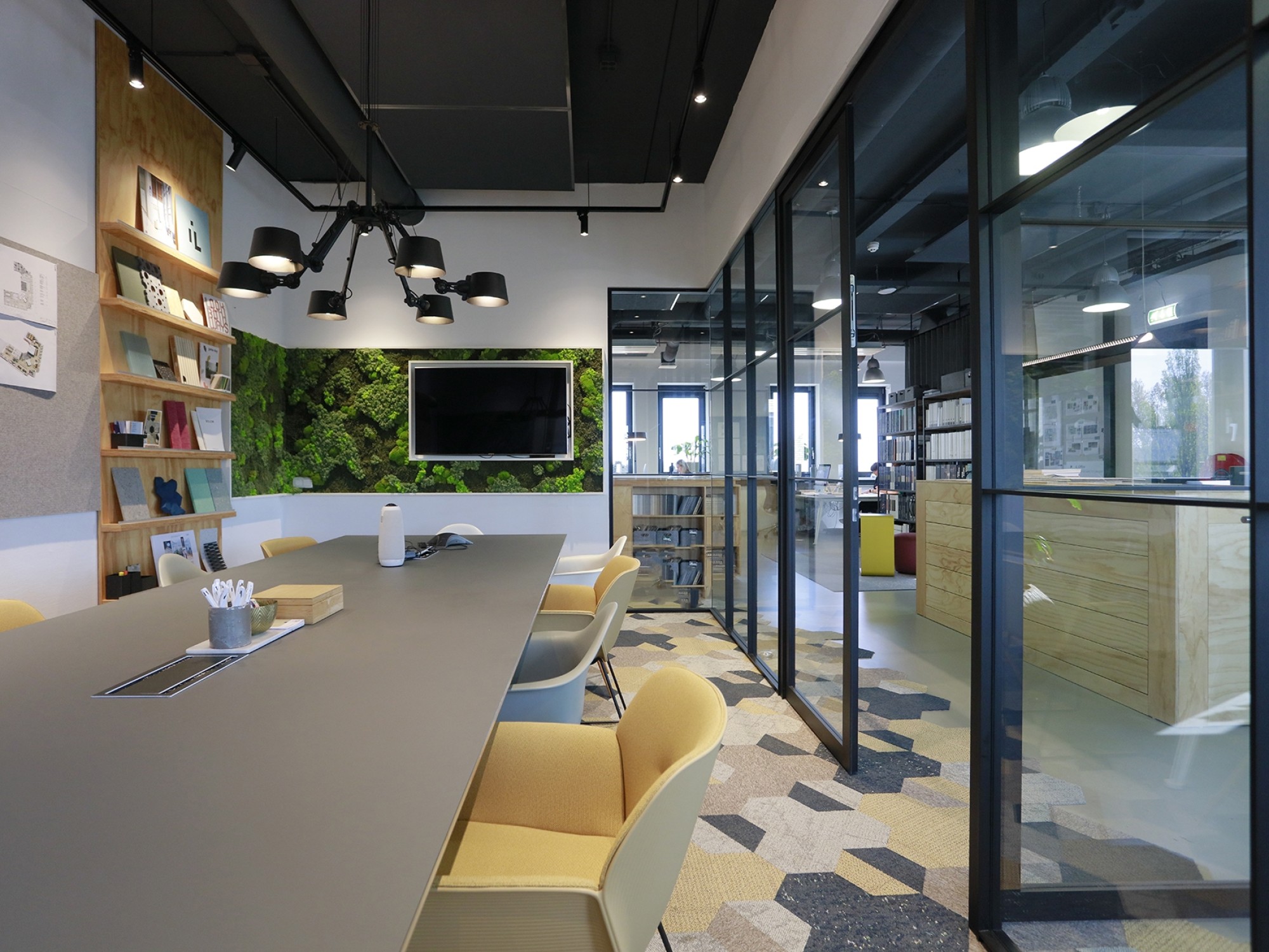 Modern meeting room with a long table and yellow chairs, glass partitions, wall-mounted shelves with books, and a television screen on the wall.