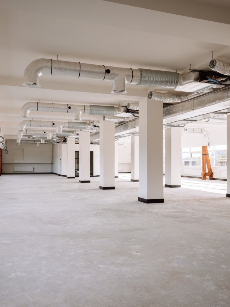 Lege kantoorruimte met betonnen vloer, witte kolommen, zichtbare ventilatiebuizen en grote ramen langs de zijkant.