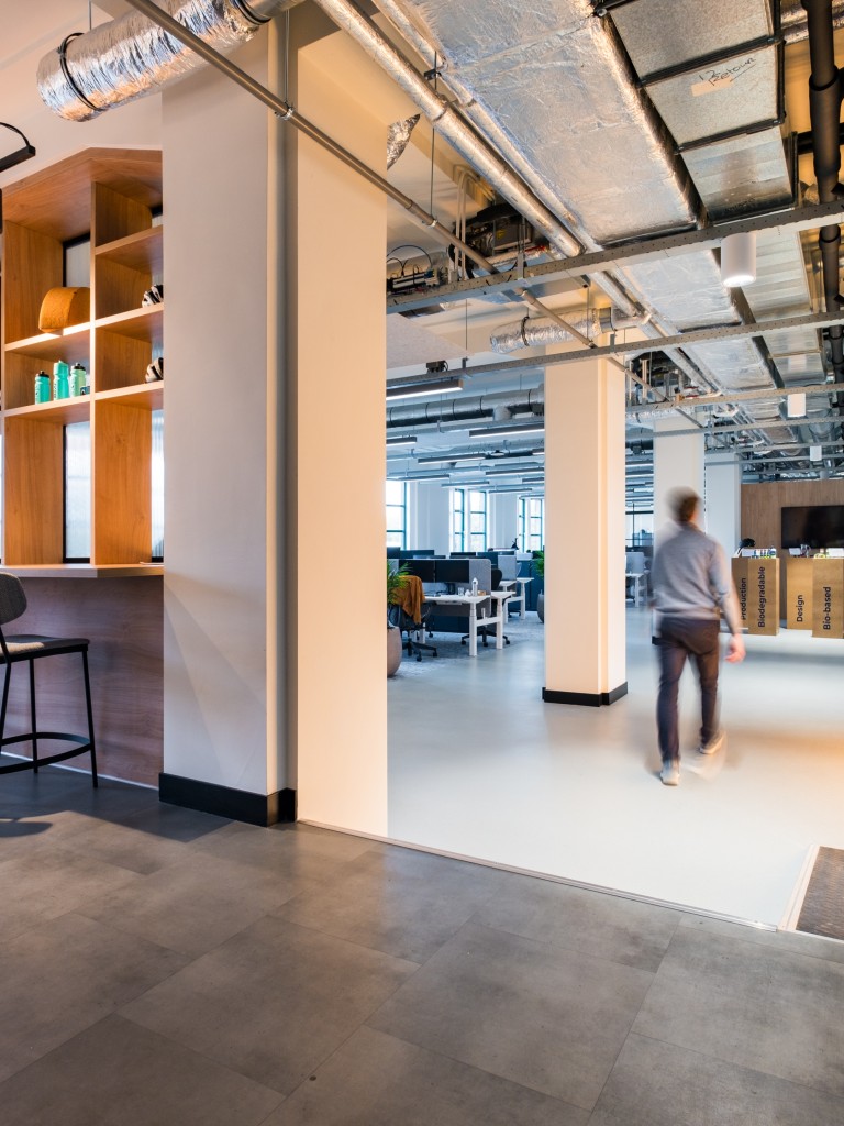 Modern kantoorinterieur met bar en hoge krukken, houten wandplanken, industriële plafonds met zichtbare leidingen en een open werkruimte met een persoon die door de ruimte loopt.
