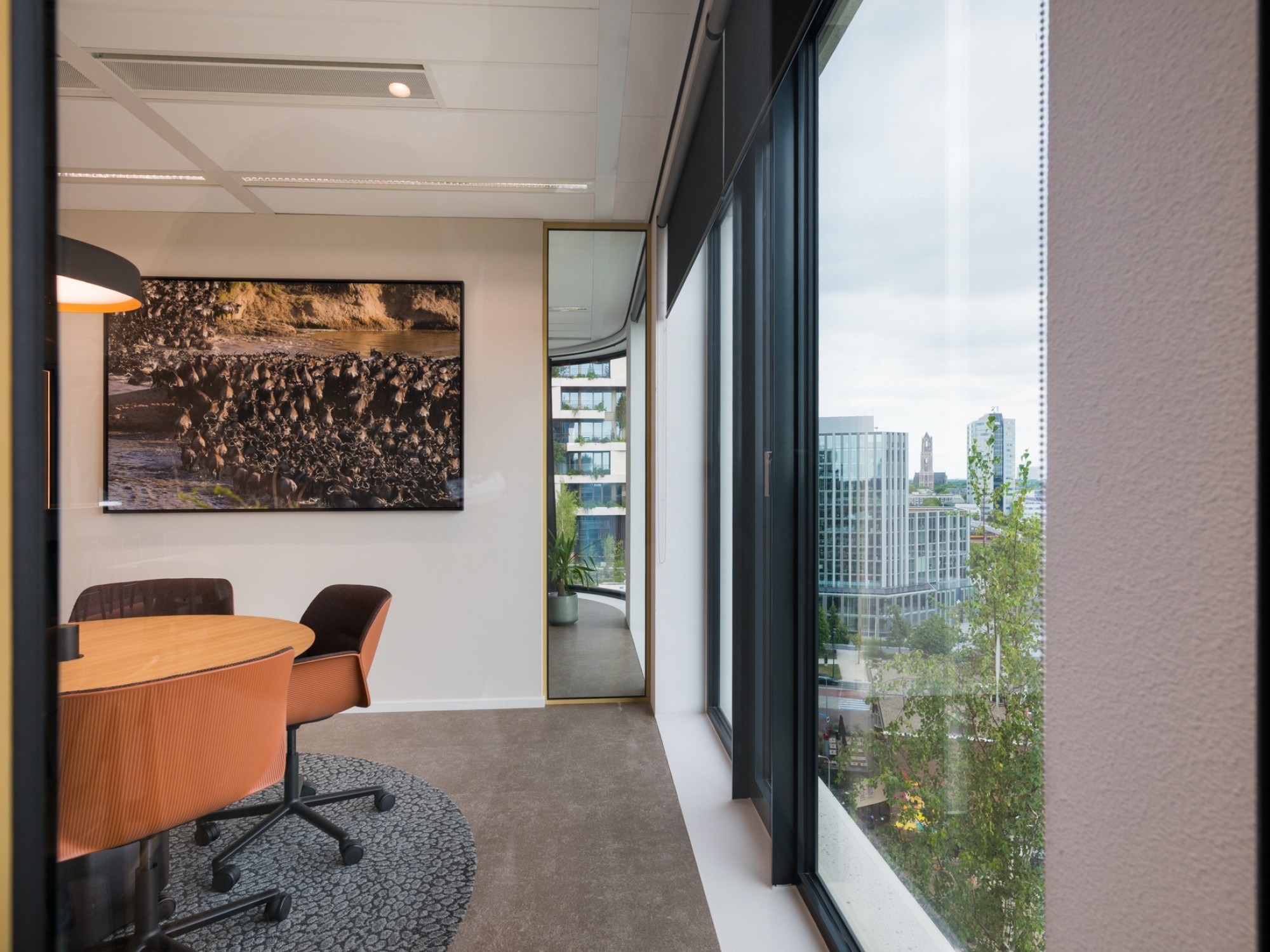 Een moderne vergaderruimte met een ronde houten tafel en bruine stoelen. Aan de muur hangt een groot kunstwerk en via de hoge ramen is uitzicht op kantoorgebouwen en groen in de stad. De ruimte is licht en strak ingericht met een rustige uitstraling.