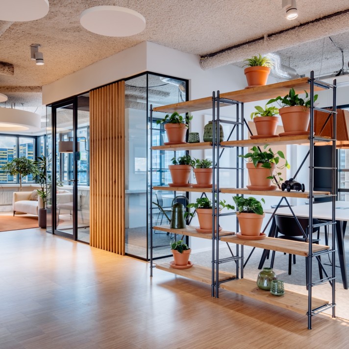 A modern office interior featuring a metal shelving unit filled with terracotta pots and green plants. In the background is a glass meeting room with black frames and wooden wall panels. The space has a light wooden floor and an industrial-style ceiling.