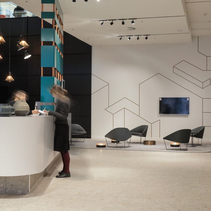 Een moderne lobby met een ronde receptiebalie waar een persoon onscherp aan staat. Rechts bevindt zich een zithoek met lage stoelen en kleine tafeltjes tegen een witte muur met minimalistische lijntekeningen. Het plafond is voorzien van spots en designver