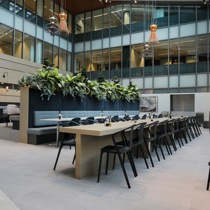 A modern office atrium with a long wooden table surrounded by black chairs. Behind the table is a raised planter filled with greenery and a built-in bench. Glass office floors overlook the open space, and decorative pendant lights hang from above.