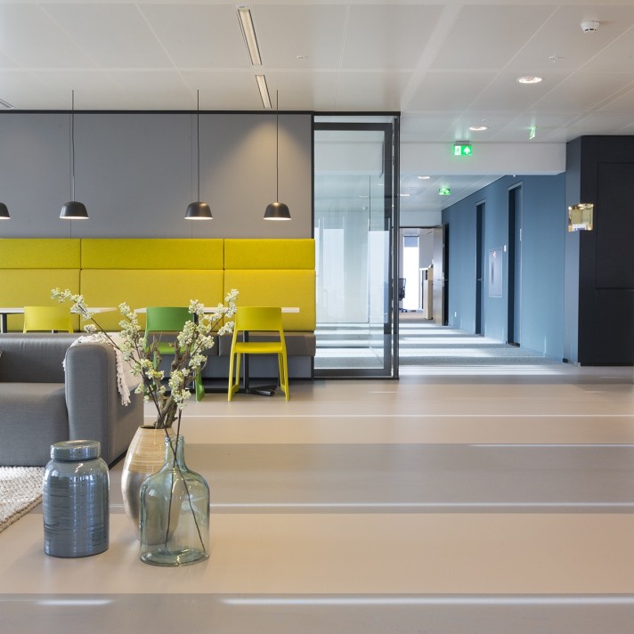 Een moderne kantoorruimte met een zithoek en een gele wandbank onder drie hanglampen. Op de voorgrond staan glazen vazen met bloemen op een tafel. Rechts loopt een gang met blauwe wanden en meerdere deuren. De ruimte heeft een lichte, strakke afwerking en