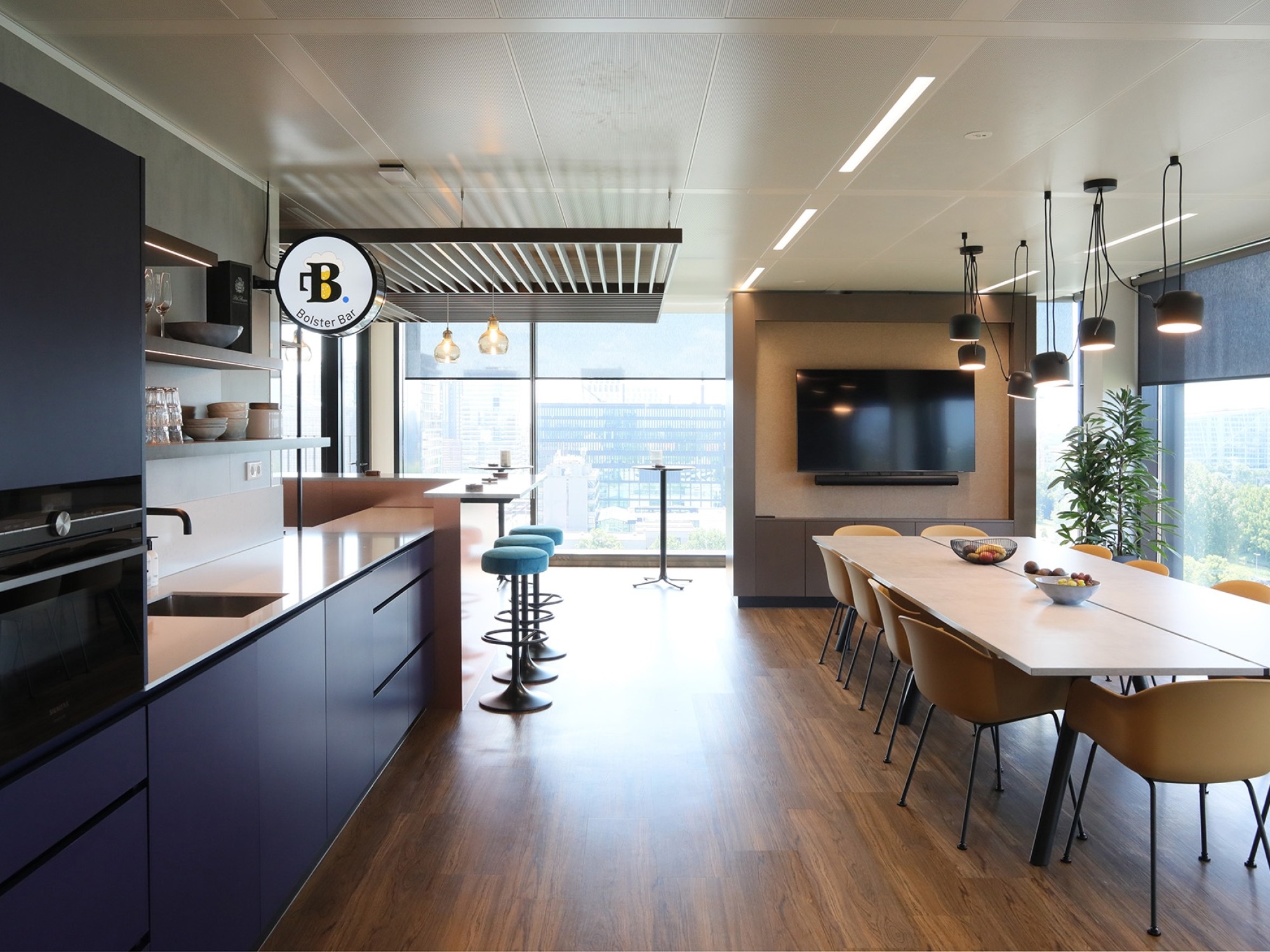 A modern office kitchen and dining area with dark cabinets and a kitchen island on the left. A long wooden dining table with chairs is positioned on the right under pendant lights. Large windows at the back let in natural light and offer a view of the cit