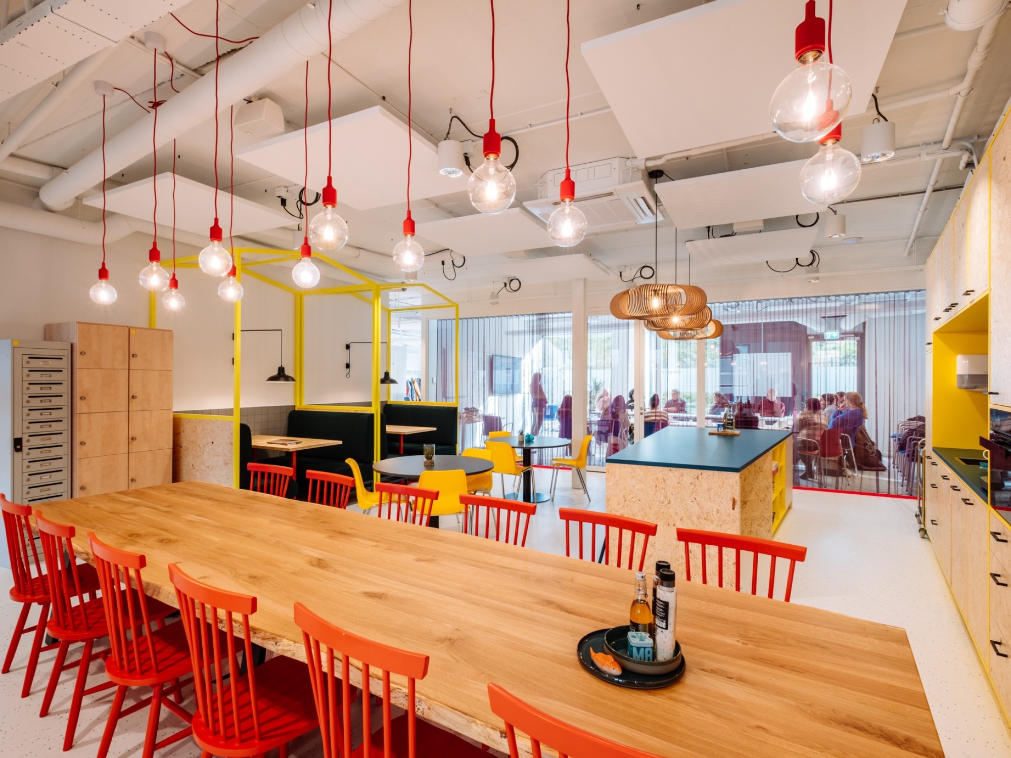 Bright and colorful office kitchen and collaborative space with a long wooden table, red chairs, hanging pendant lights, yellow accents, and a glass-walled meeting room in the background.