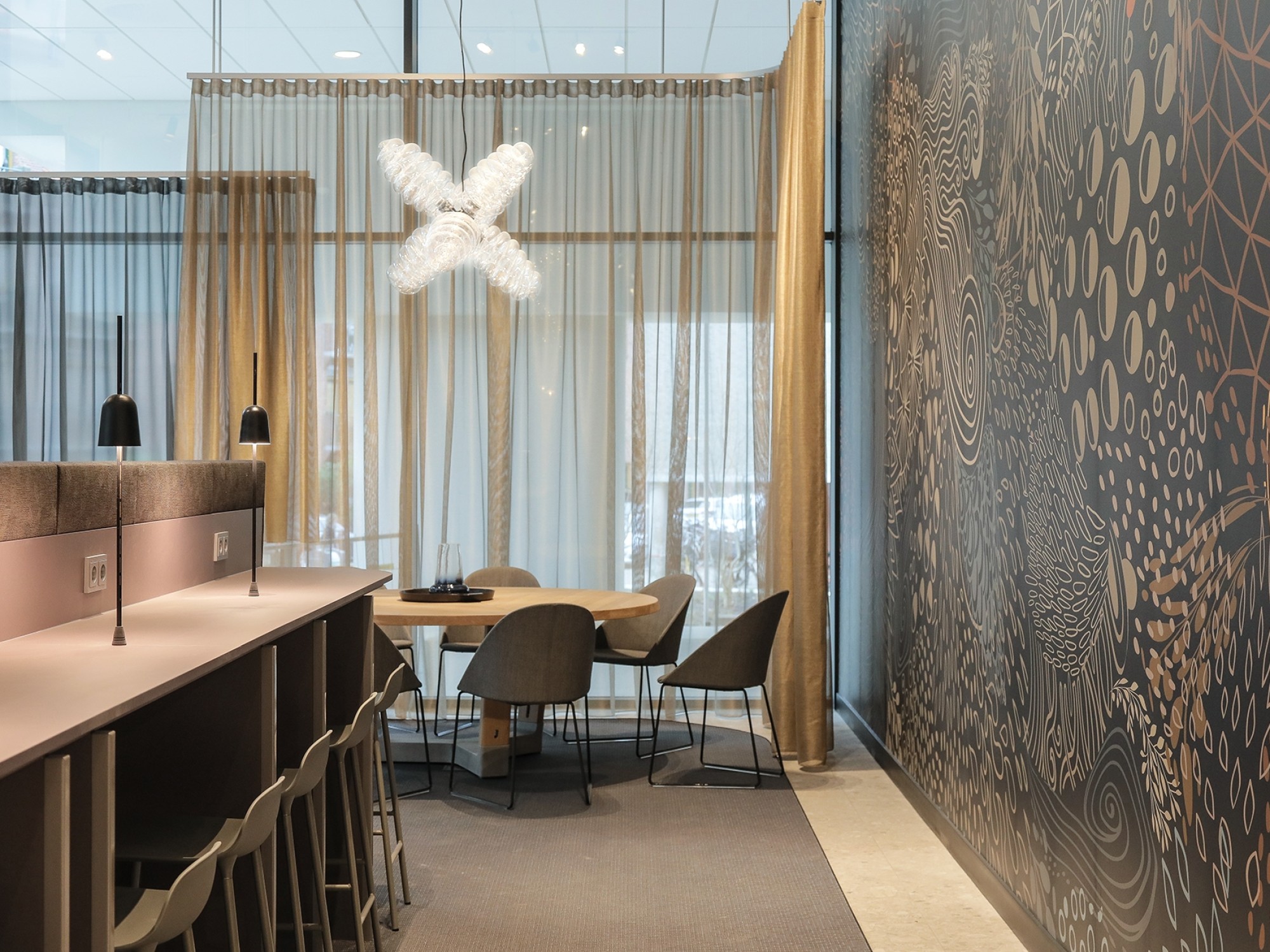 Een moderne vergaderruimte met een hoge bartafel en krukken aan de linkerzijde. Achterin staat een ronde tafel met stoelen bij grote ramen met transparante gordijnen. Aan de rechterkant is een wand met een decoratief patroon zichtbaar en boven de tafel ha