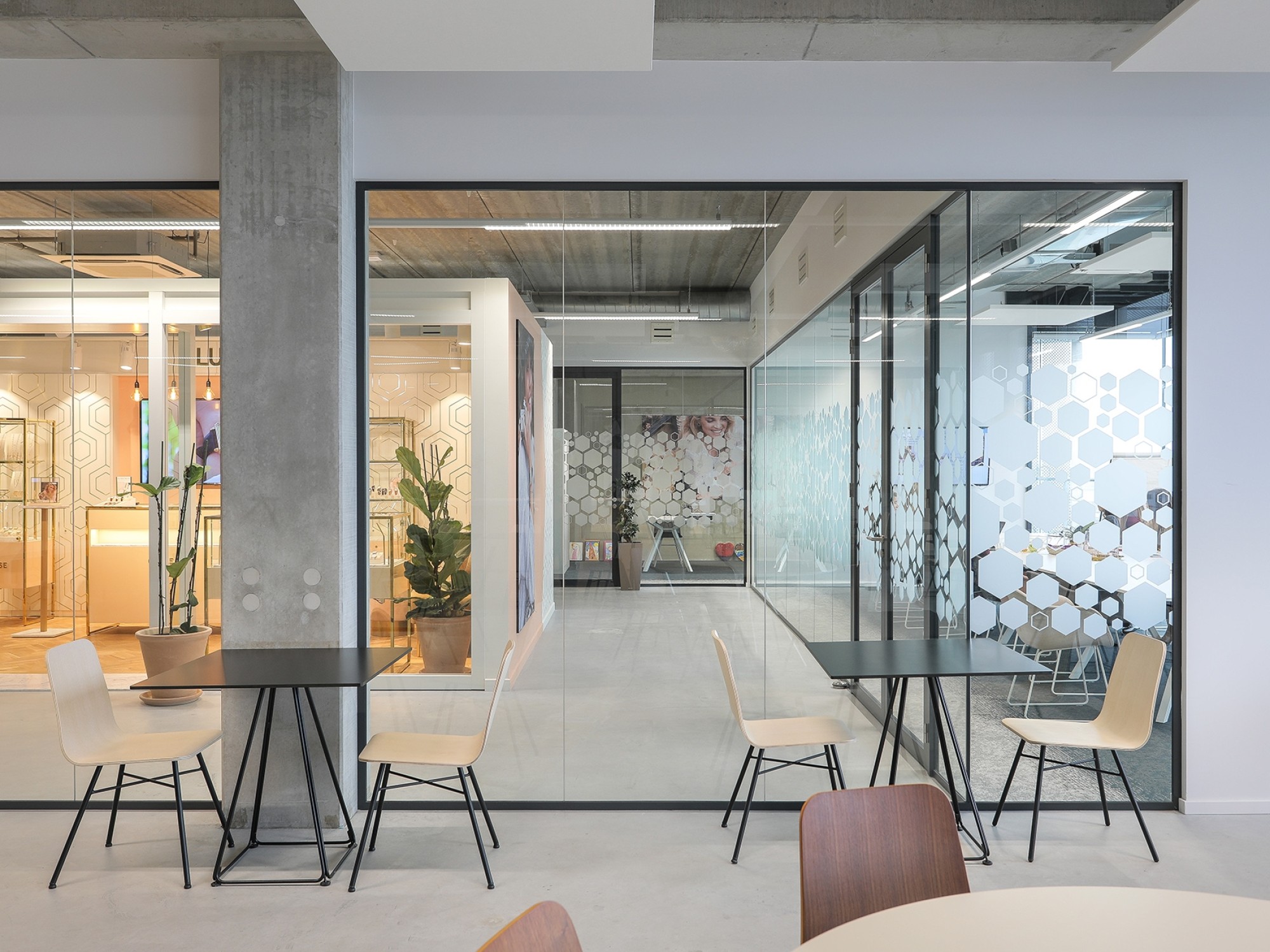 Een moderne kantoorruimte met glazen vergaderruimtes en zwarte frames. Op het glas zijn subtiele patronen zichtbaar. Voor de glazen wanden staan kleine ronde tafels met lichte stoelen. De ruimte heeft een industriële uitstraling met betonnen kolommen en e