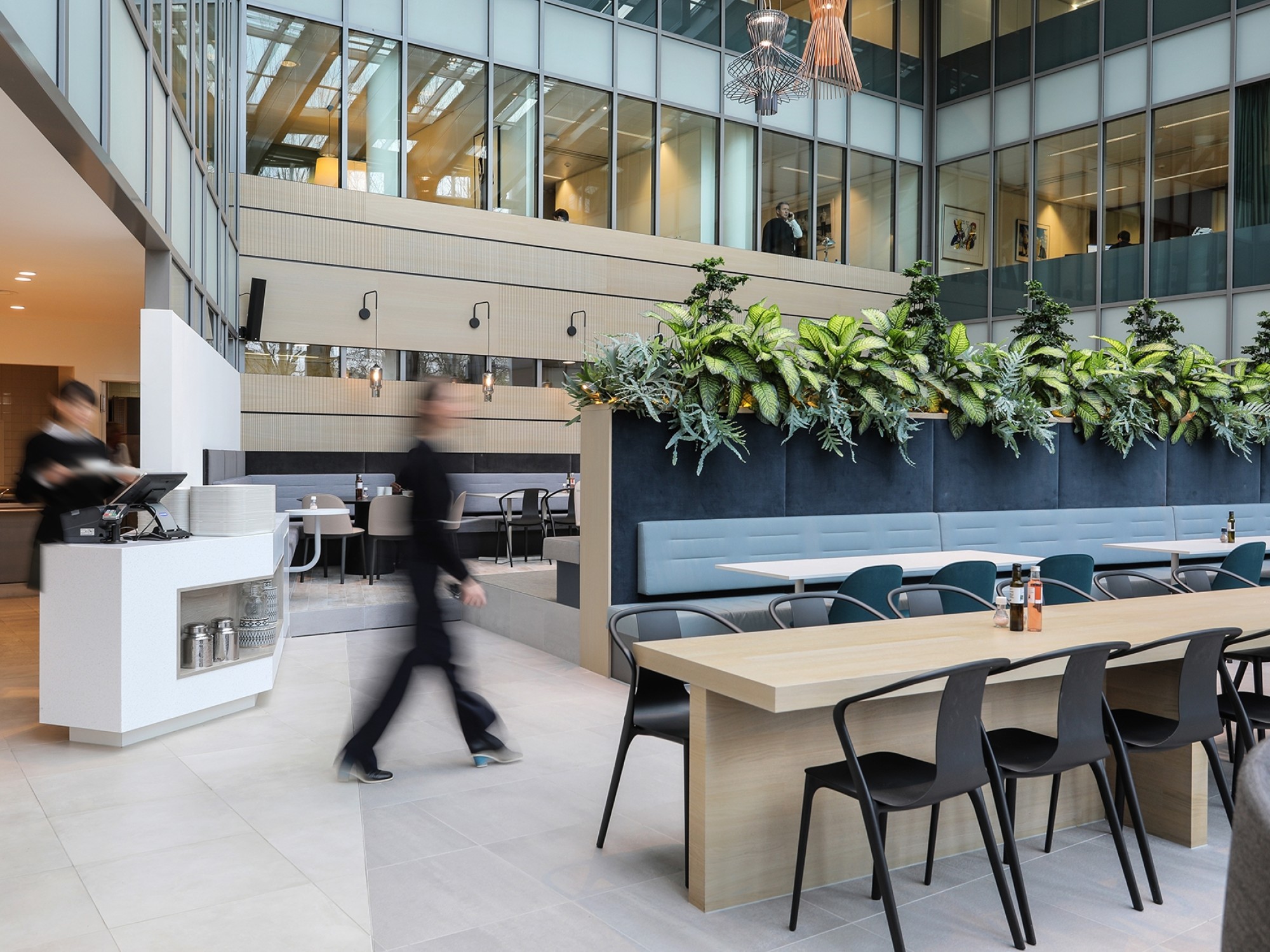 A modern office atrium with a long wooden table and black chairs. A raised planter with greenery and a built-in bench runs behind the table. On the left, a coffee counter is visible while a person walks past, slightly blurred. Glass office levels overlook