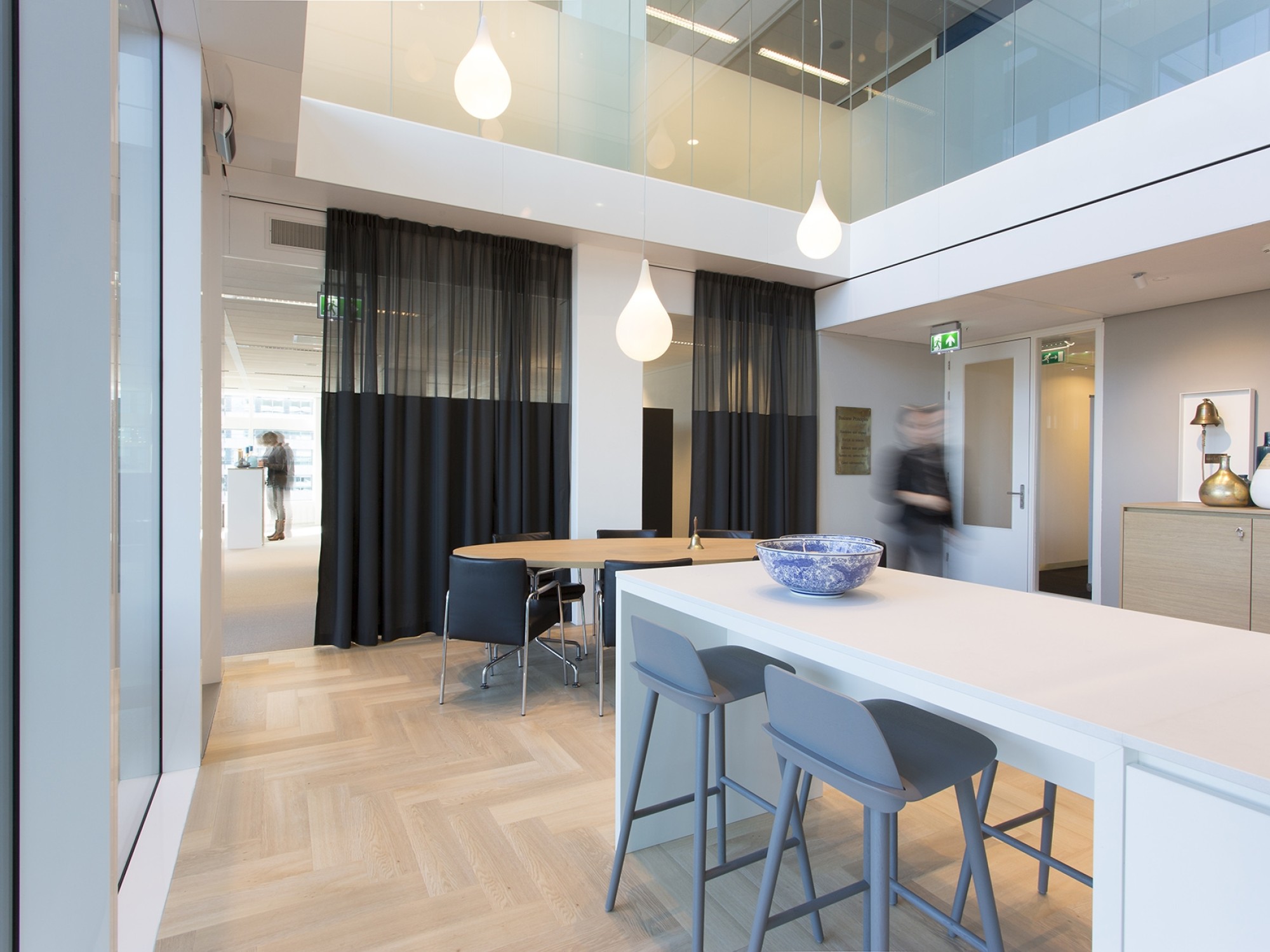 Een moderne kantoorruimte met een hoge vide en lichte houten visgraatvloer. Op de voorgrond staat een wit kookeiland met barkrukken en een schaal erop. Achterin bevindt zich een vergaderruimte met een tafel en stoelen, deels afgescheiden door donkere gord