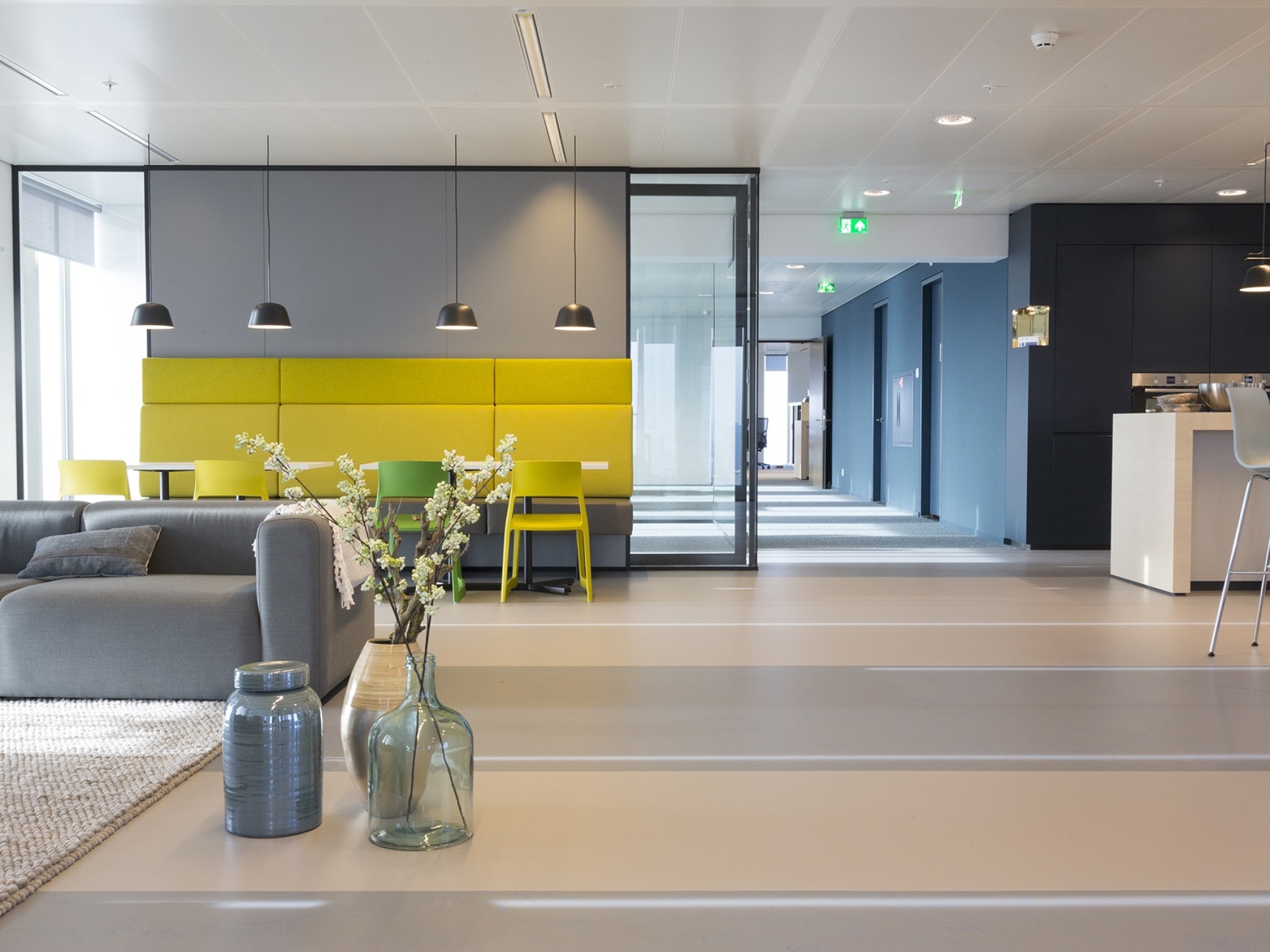 Een moderne kantoorruimte met een zithoek en een gele wandbank onder drie hanglampen. Op de voorgrond staan glazen vazen met bloemen op een tafel. Rechts loopt een gang met blauwe wanden en meerdere deuren. De ruimte heeft een lichte, strakke afwerking en