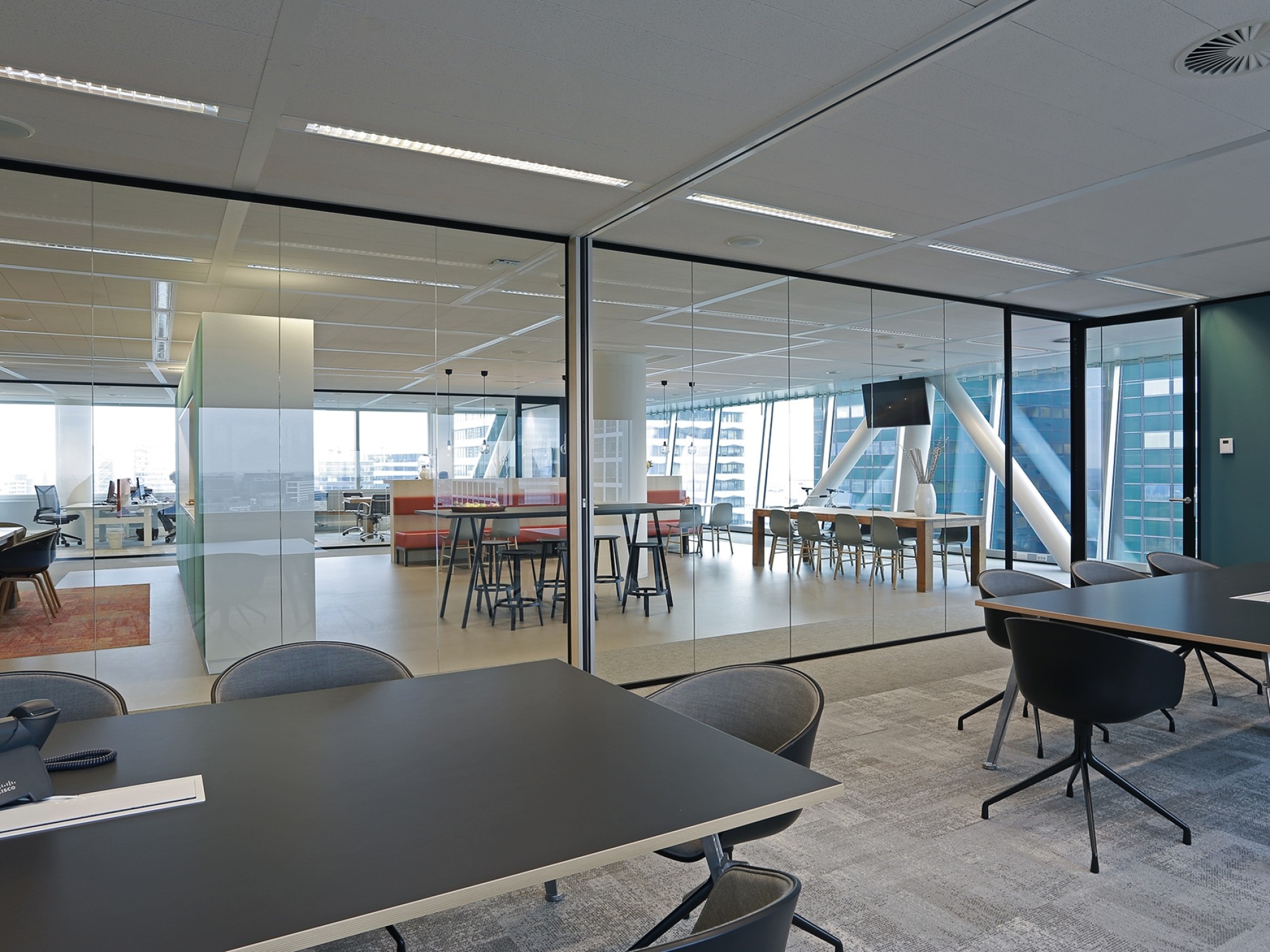 Een moderne kantoorruimte met zwarte tafels en stoelen op de voorgrond. Achter glazen wanden is een open werk- en ontmoetingsruimte zichtbaar met hoge tafels en grote ramen. De ruimte heeft een strakke afwerking en veel daglicht.