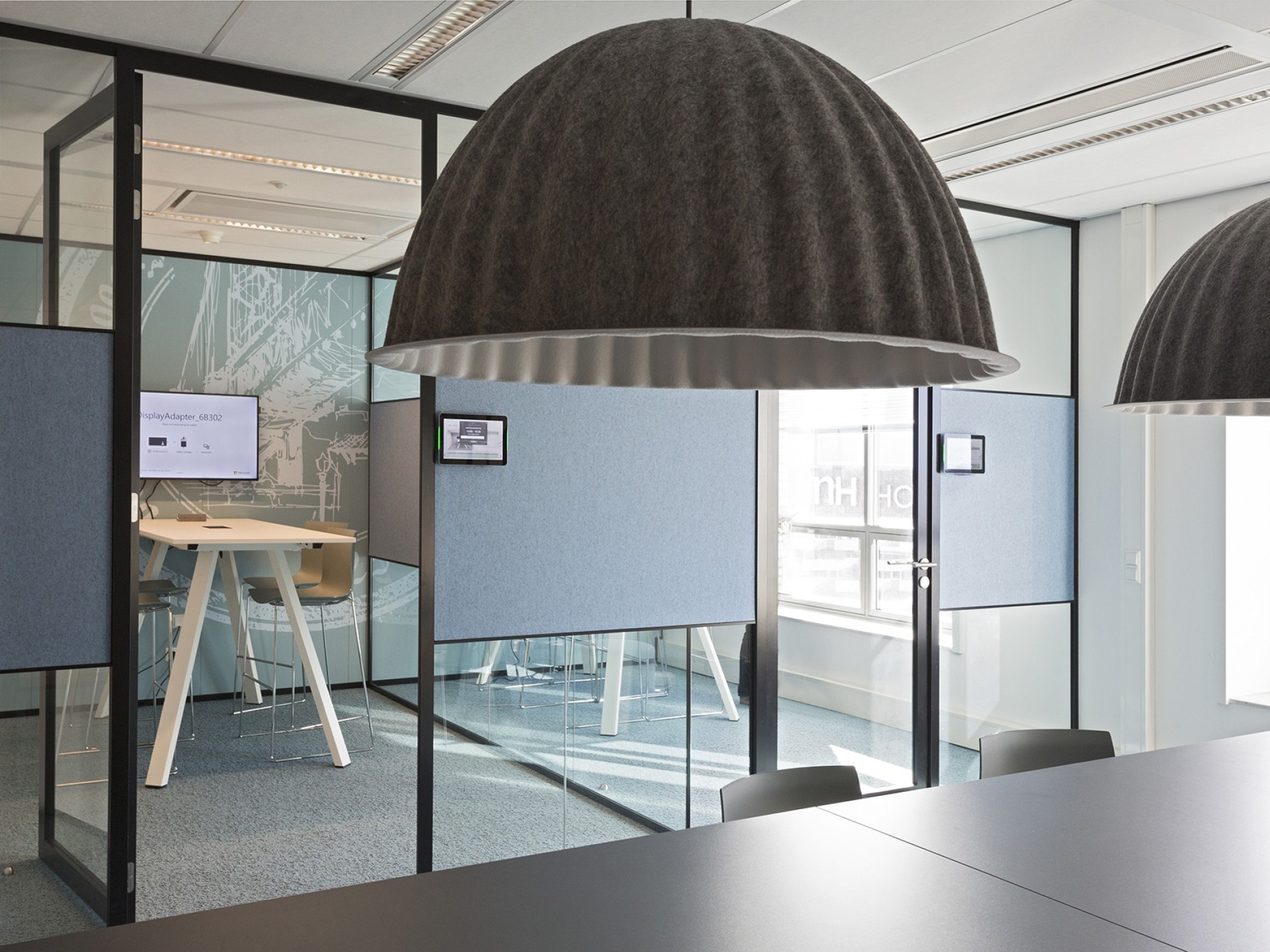 A modern office interior with glass meeting rooms framed in black. Two large dark pendant lights hang in the foreground above a table. Inside the meeting rooms are high tables, chairs, and blue privacy panels, with large windows allowing natural light.