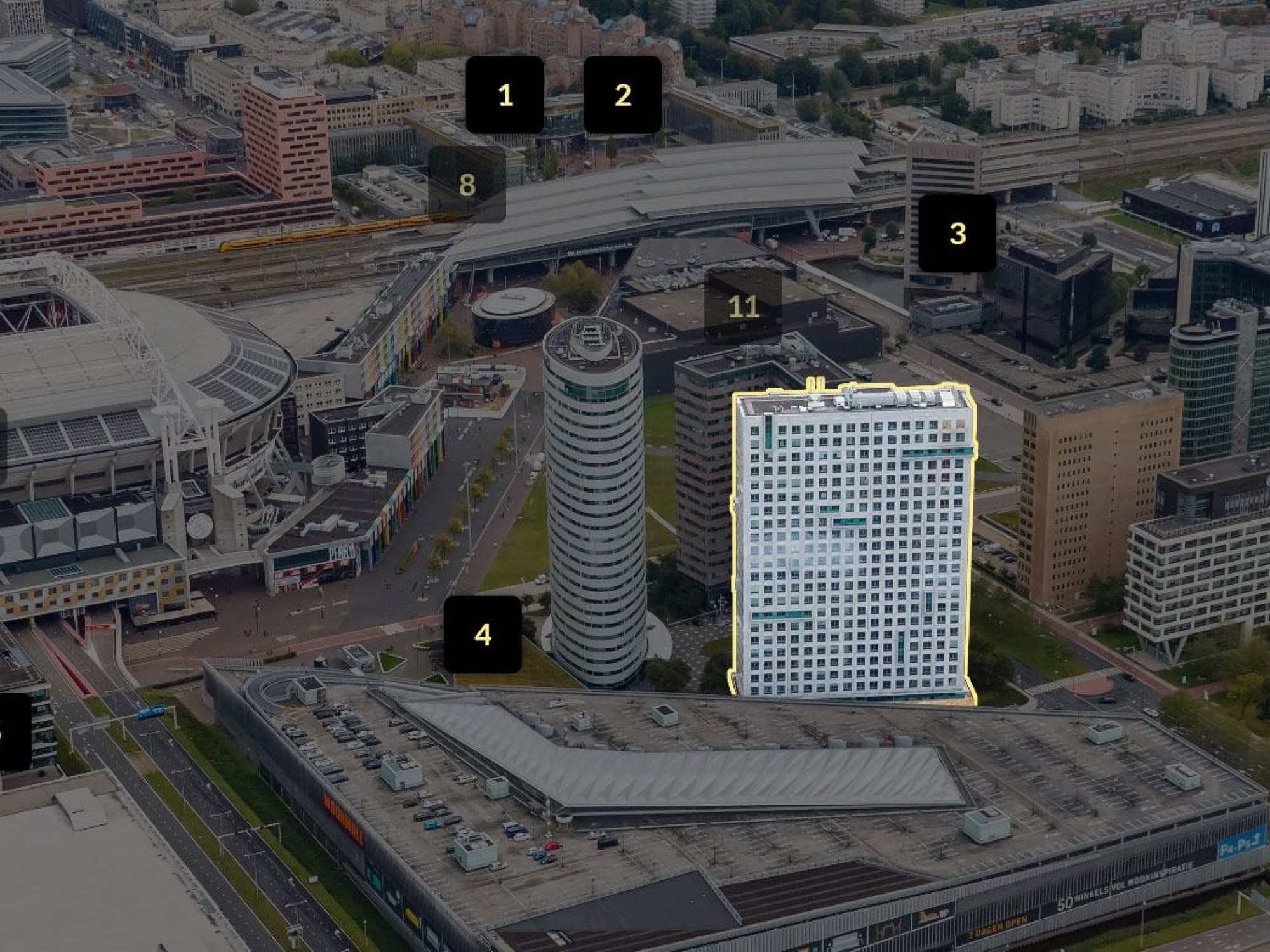 Een luchtfoto van een stedelijk gebied met meerdere grote gebouwen en wegen. Eén hoog, rechthoekig gebouw is met een lichte omlijning gemarkeerd. Op verschillende locaties in de afbeelding staan zwarte labels met witte cijfers die specifieke gebouwen aand
