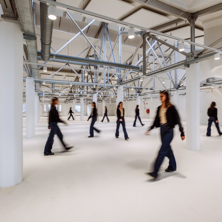 Een grote, open industriële ruimte met witte kolommen en zichtbare stalen constructies en ventilatiebuizen aan het plafond. Meerdere personen lopen door de ruimte, deels onscherp door beweging. De vloer is licht en de ruimte oogt ruim en minimalistisch.