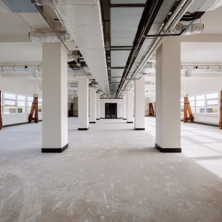 Een grote, lege kantoorruimte met betonnen vloer en witte kolommen in een rij. Aan het plafond zijn ventilatiekanalen en leidingen zichtbaar. Grote ramen aan beide zijden zorgen voor veel daglicht in de open ruimte.