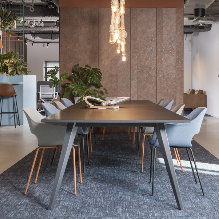 Een moderne vergaderruimte met een grote donkere tafel en grijze stoelen eromheen. Boven de tafel hangt een designlamp met zichtbare gloeilampen. Op de achtergrond zijn planten en een industriële kantooromgeving zichtbaar met een open plafond en metalen d