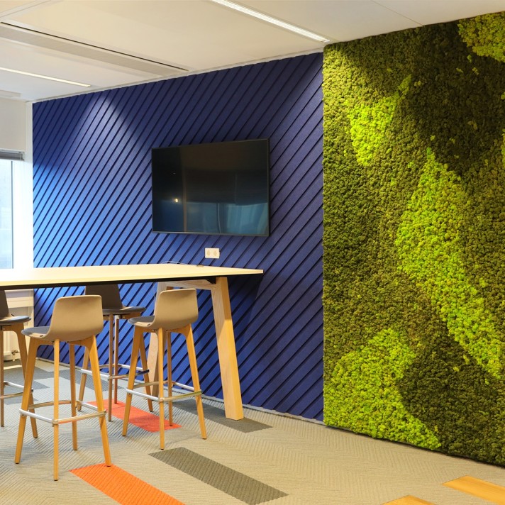 Een moderne kantoorruimte met een hoge tafel en houten barkrukken tegen een blauwe wand met diagonale latten. Aan de muur hangt een flatscreen. Rechts bevindt zich een groene plantenwand met een opvallend patroon. De vloer heeft kleurrijke accenten in ora
