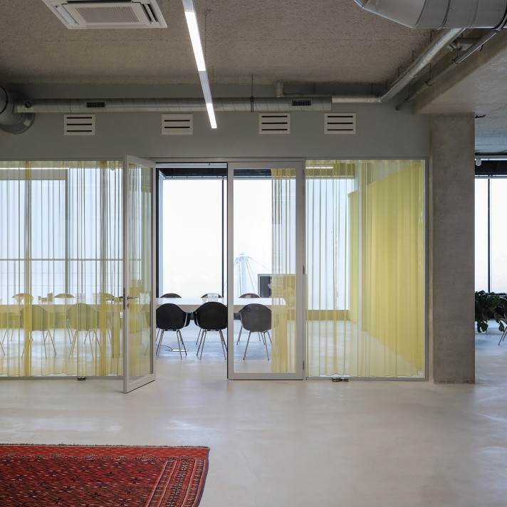 Een moderne kantoorruimte met een glazen vergaderruimte achter gele transparante gordijnen. Binnen staan een tafel en meerdere stoelen. De ruimte heeft een industriële uitstraling met betonnen kolommen, zichtbare ventilatiebuizen en een strakke vloer.
