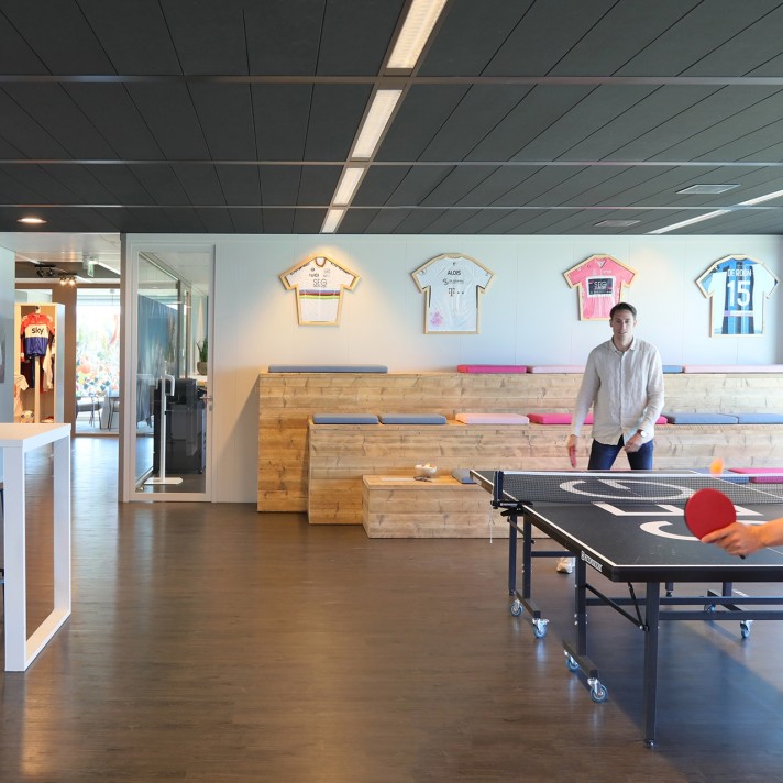 Een moderne kantoorruimte met een tafeltennistafel op de voorgrond waar twee personen aan spelen. Aan de muur hangen ingelijste sportshirts boven een houten tribune met zitplaatsen. De ruimte heeft een donkere vloer en een strak plafond met verlichting.
