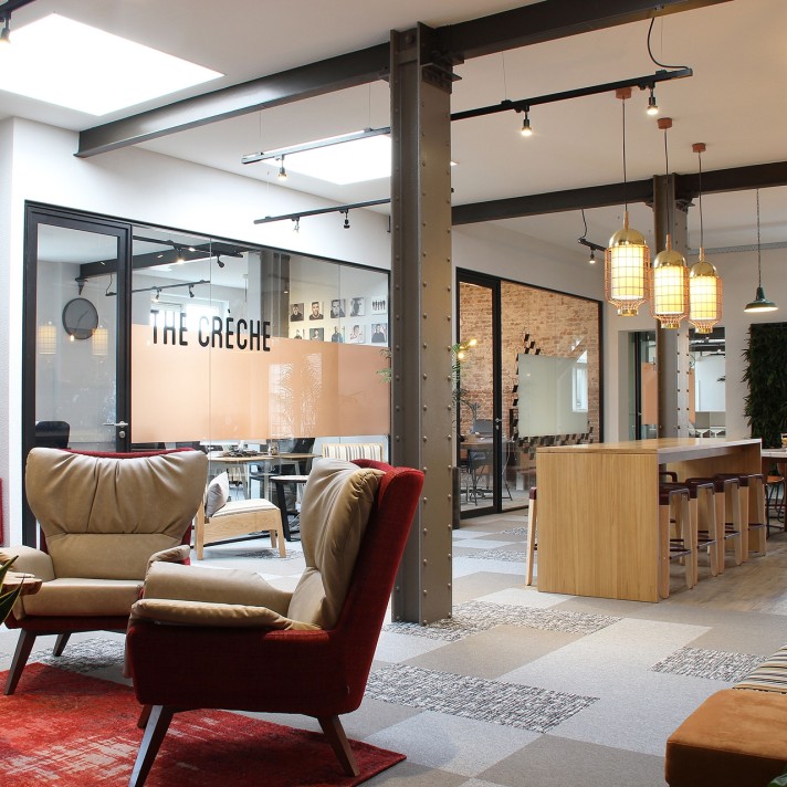 Een moderne kantoorruimte met een gezellige zithoek met fauteuils op de voorgrond. Achter glazen wanden is een vergaderruimte zichtbaar met de tekst “The Crèche” op het glas. Rechts bevindt zich een houten bar met hanglampen en een open werkruimte met ind