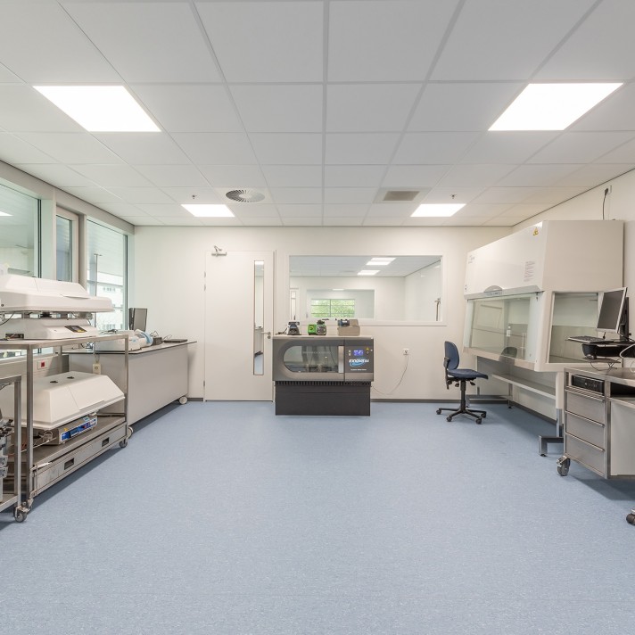 Een schone, witte laboratoriumruimte met werkbanken en apparatuur aan beide zijden. In het midden staat een gekoelde opslagunit en rechts een werkstation met stoel en veiligheidskast. De ruimte heeft een blauwe vloer en helder plafondlicht.