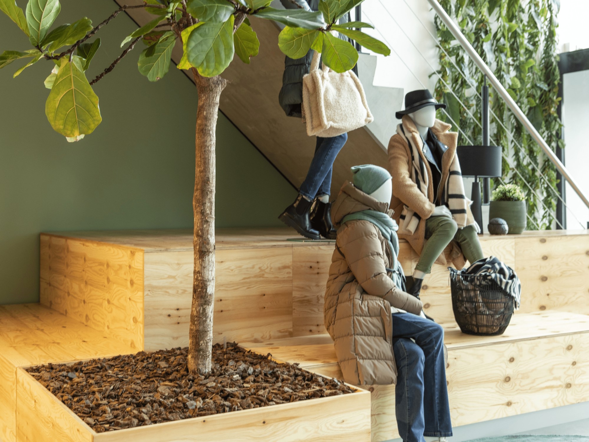 Een moderne binnenruimte met houten zitblokken rond een boom in een plantenbak. Twee personen zitten op de houten constructie, gekleed in winterjassen en mutsen, terwijl een derde persoon langsloopt. De ruimte heeft een groene wand en een lichte, open uit