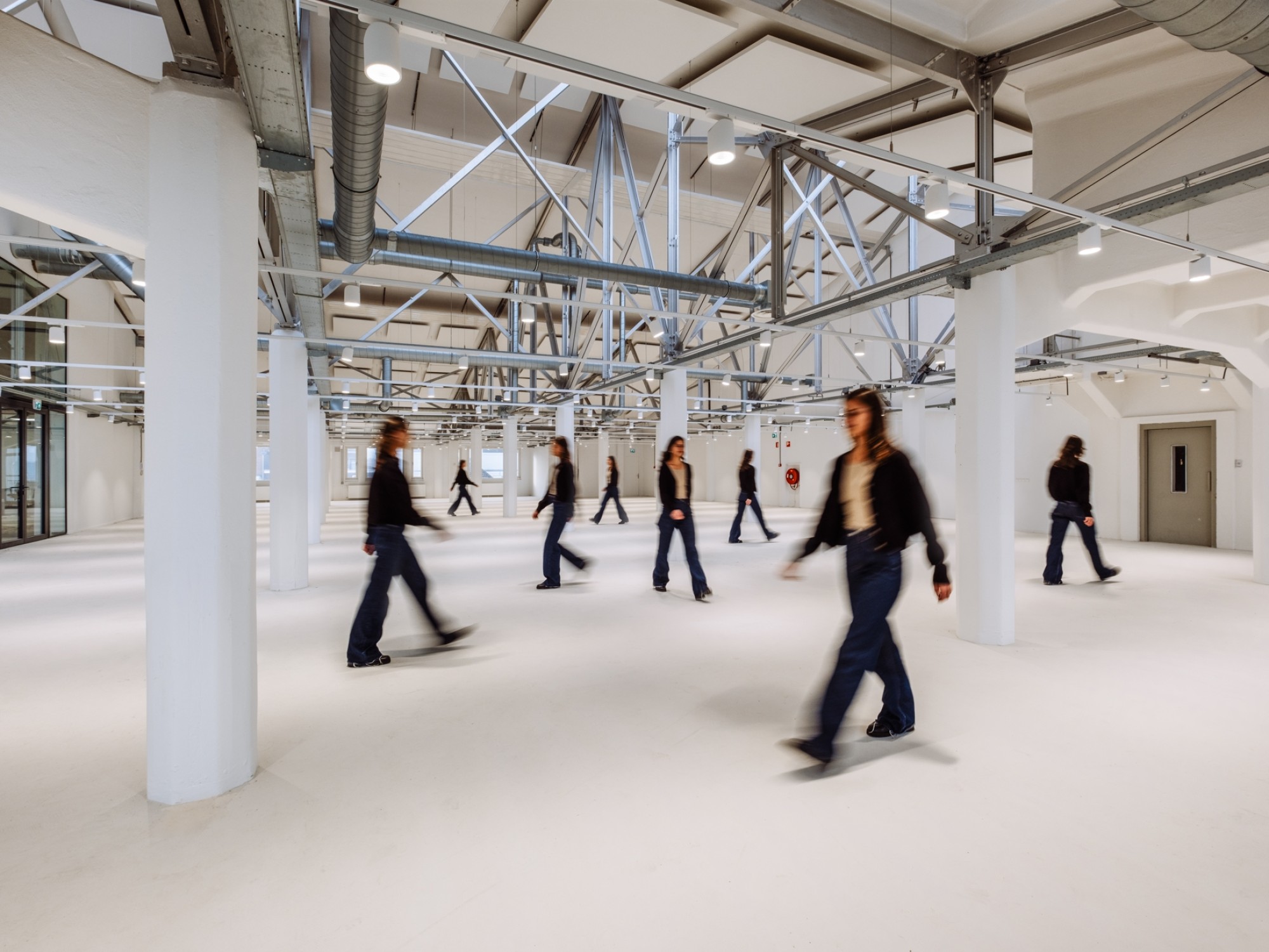 Een grote, open industriële ruimte met witte kolommen en zichtbare stalen constructies en ventilatiebuizen aan het plafond. Meerdere personen lopen door de ruimte, deels onscherp door beweging. De vloer is licht en de ruimte oogt ruim en minimalistisch.