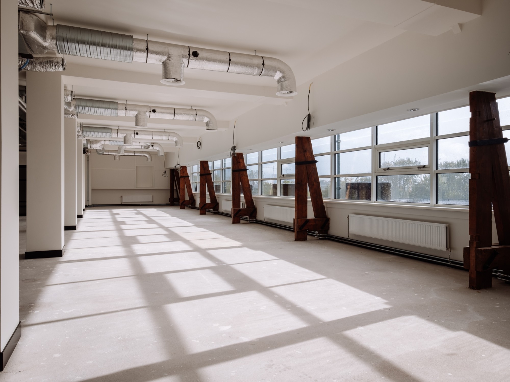 Een ruime, lege kantoorruimte met grote ramen langs één zijde waardoor veel daglicht naar binnen valt. De vloer is onafgewerkt en langs de ramen staan houten steunen. Aan het plafond zijn ventilatiekanalen en leidingen zichtbaar, wat de ruimte een industr