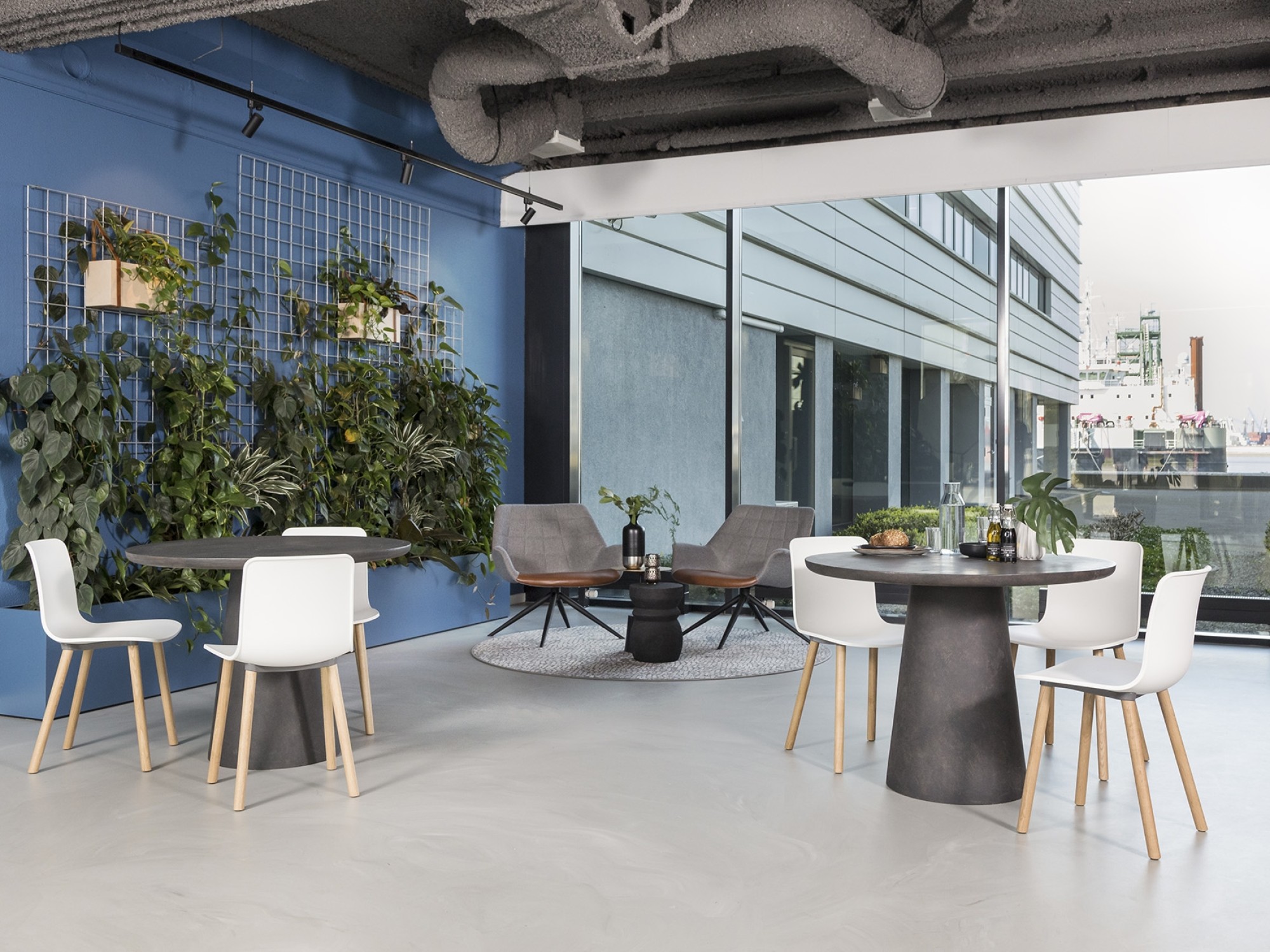Een moderne kantoorruimte met ronde tafels en witte stoelen bij grote ramen met uitzicht naar buiten. Links staat een blauwe wand met klimplanten en groene beplanting. De ruimte heeft een open, industriële uitstraling met zichtbare ventilatiebuizen aan he