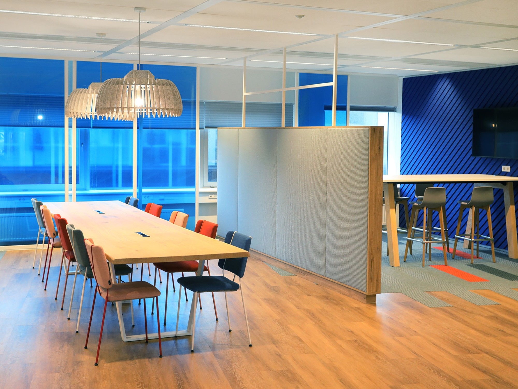 Een moderne kantoorruimte met een lange houten vergadertafel en gekleurde stoelen op een lichte houten vloer. Boven de tafel hangt een grote designlamp. Rechts bevindt zich een hoge tafel met barkrukken tegen een blauwe wand met een flatscreen. Grote rame