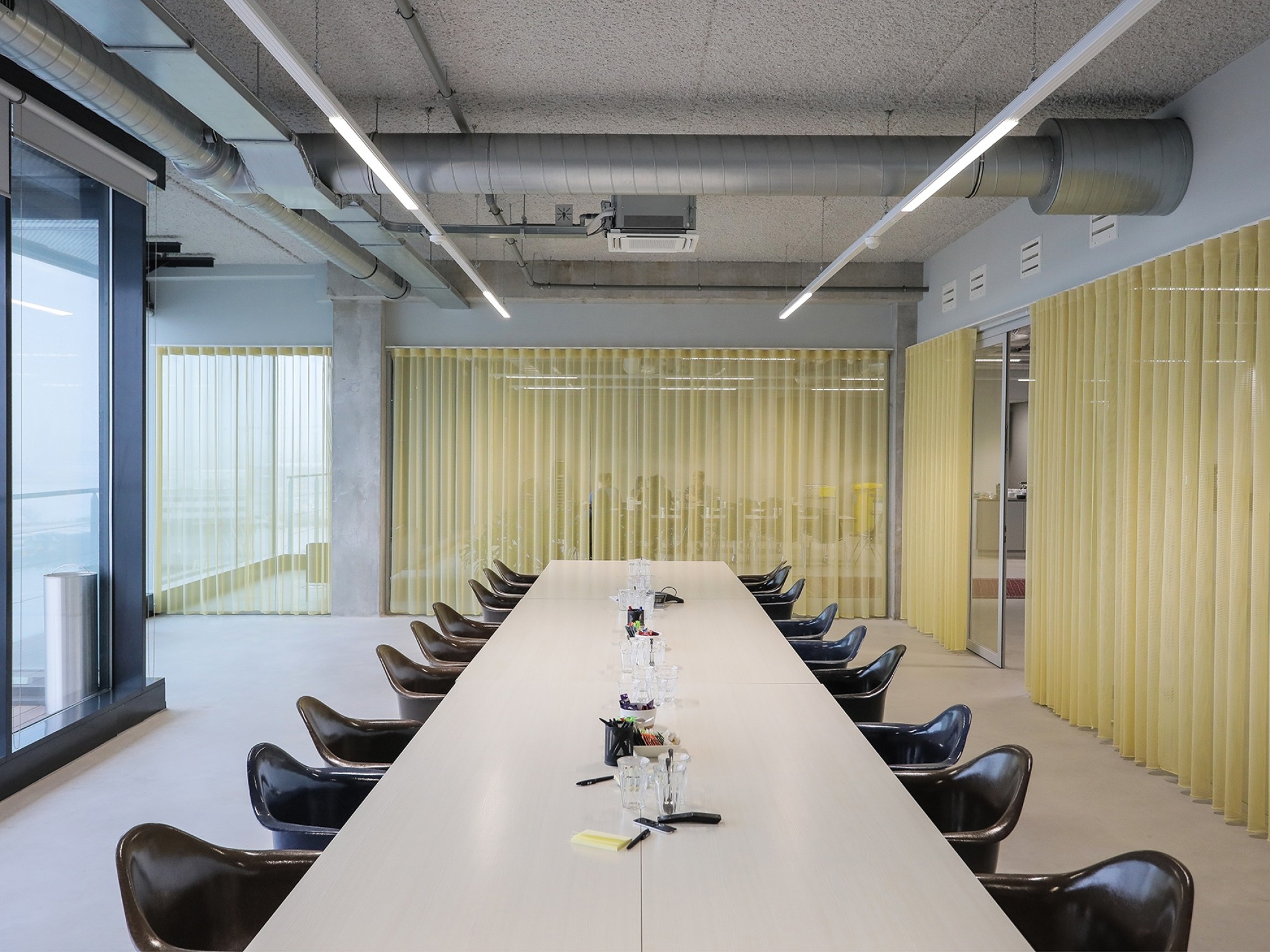 Een moderne vergaderruimte met een lange witte tafel en zwarte stoelen aan beide zijden. Op tafel staan kleine microfoons en notitieblokken. De ruimte is omgeven door glazen wanden met gele transparante gordijnen en heeft een industrieel plafond met zicht