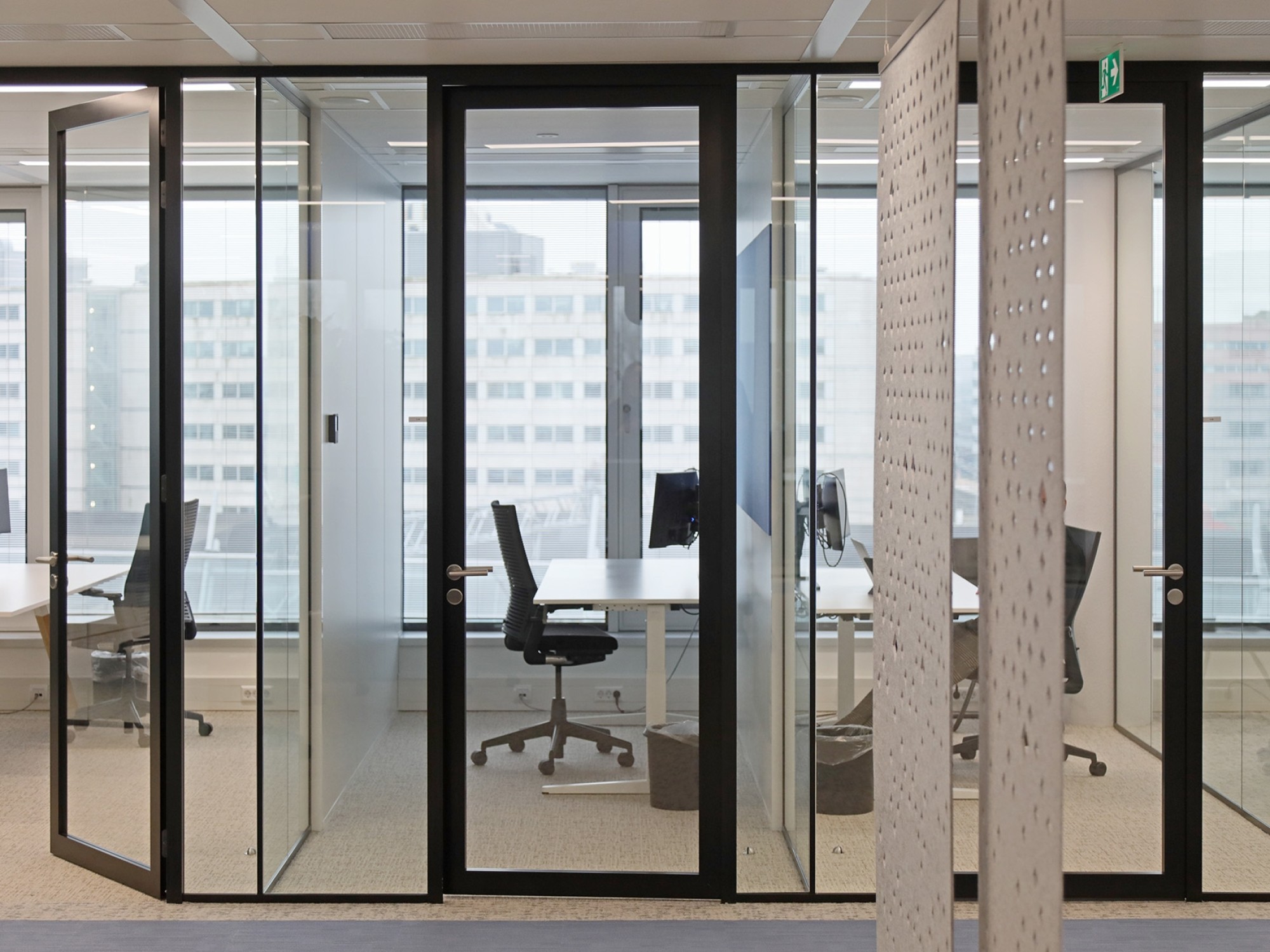 Een moderne kantoorruimte met glazen vergader- en werkruimtes met zwarte frames. Binnen staan bureaus met bureaustoelen bij grote ramen met uitzicht op andere gebouwen. Op de voorgrond is een houten wand met een geperforeerd patroon zichtbaar.