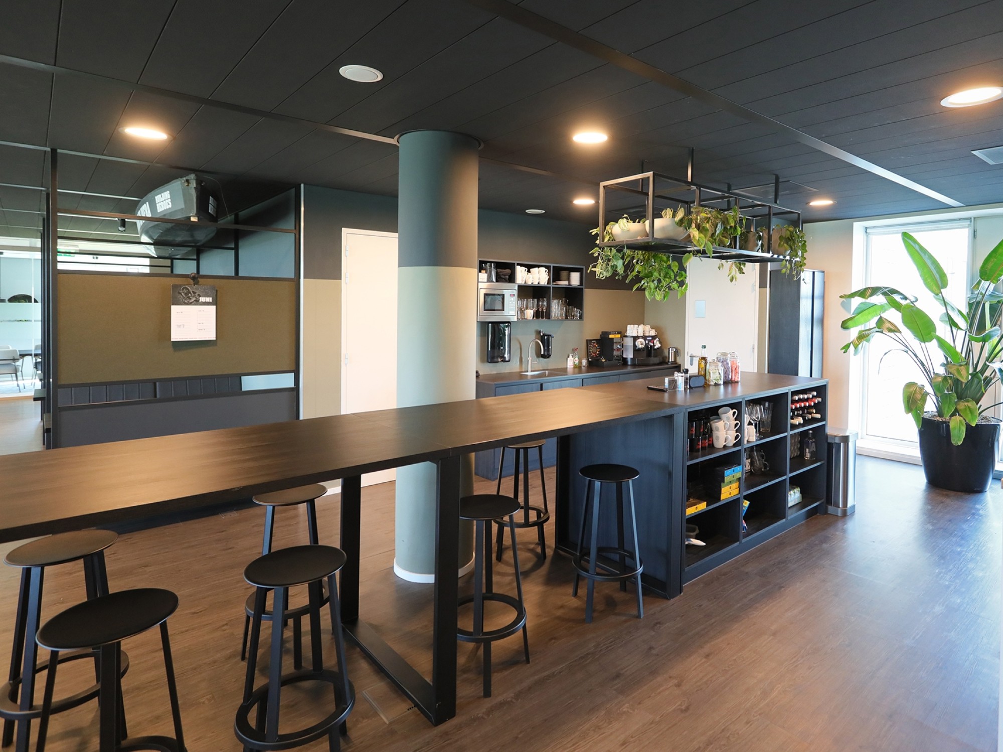 Een moderne kantoorpantry met een lange hoge tafel en zwarte barkrukken. Achter de bar staan koffiemachines, glazen en flessen op open planken. Planten hangen boven de bar en grote ramen zorgen voor veel natuurlijk licht in de ruimte.