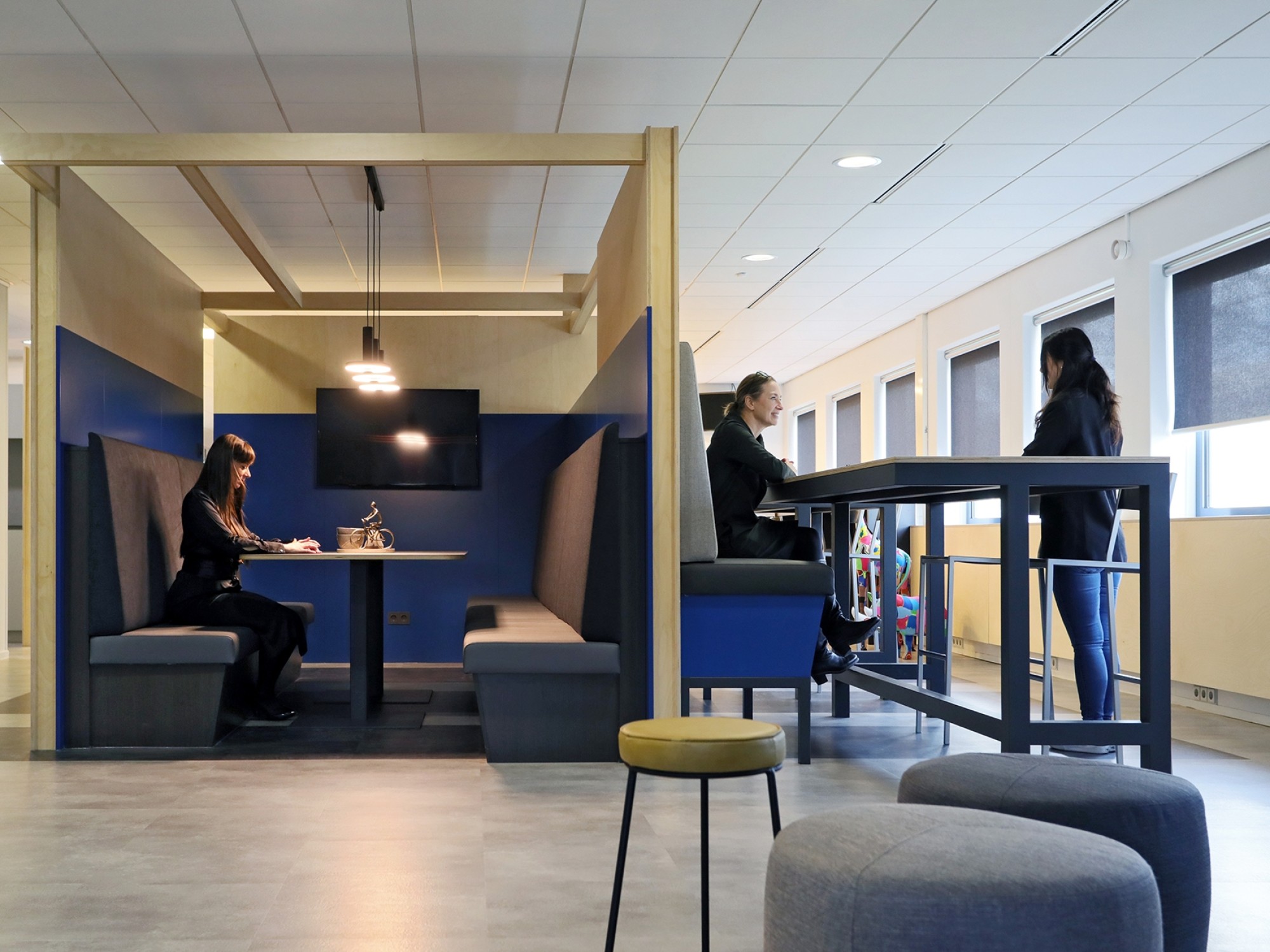 Een moderne kantoorruimte met twee verschillende overlegplekken. Links zit een persoon in een afgesloten zithoek met blauwe wanden aan een tafel. Rechts staan twee personen aan een hoge tafel met barkrukken bij een raam. Op de voorgrond staan ronde poefje