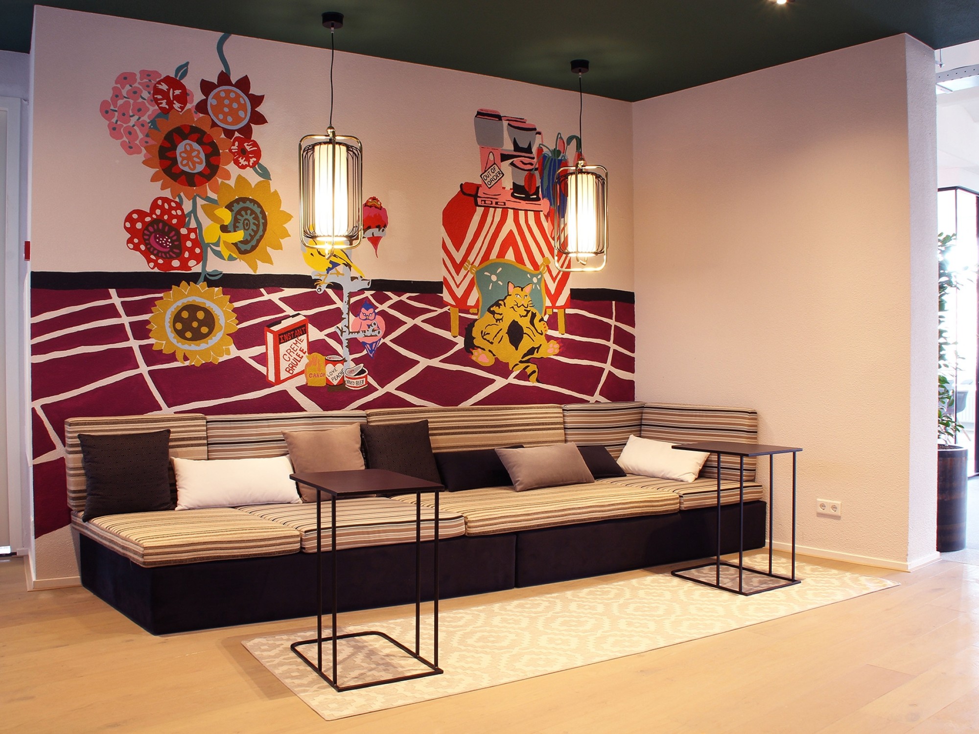 Een moderne zithoek met een lange bank met kussens en twee zwarte bijzettafels op een licht vloerkleed. Aan de muur hangt een kleurrijke muurschildering met tandwielen en illustraties. Boven de bank hangen twee hanglampen en de ruimte heeft een warme, cre