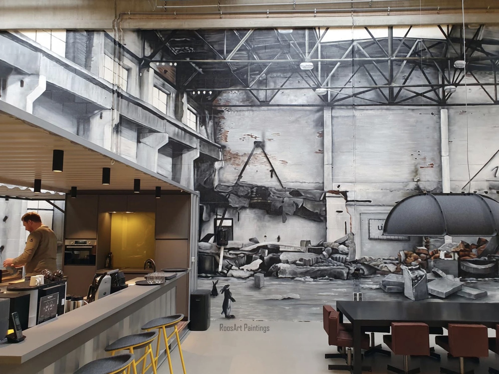 A spacious industrial-style office interior with high ceilings and exposed steel beams. A large black-and-white mural covers the back wall. On the left, a bar counter with stools and a person working behind it is visible, while tables and chairs are arran