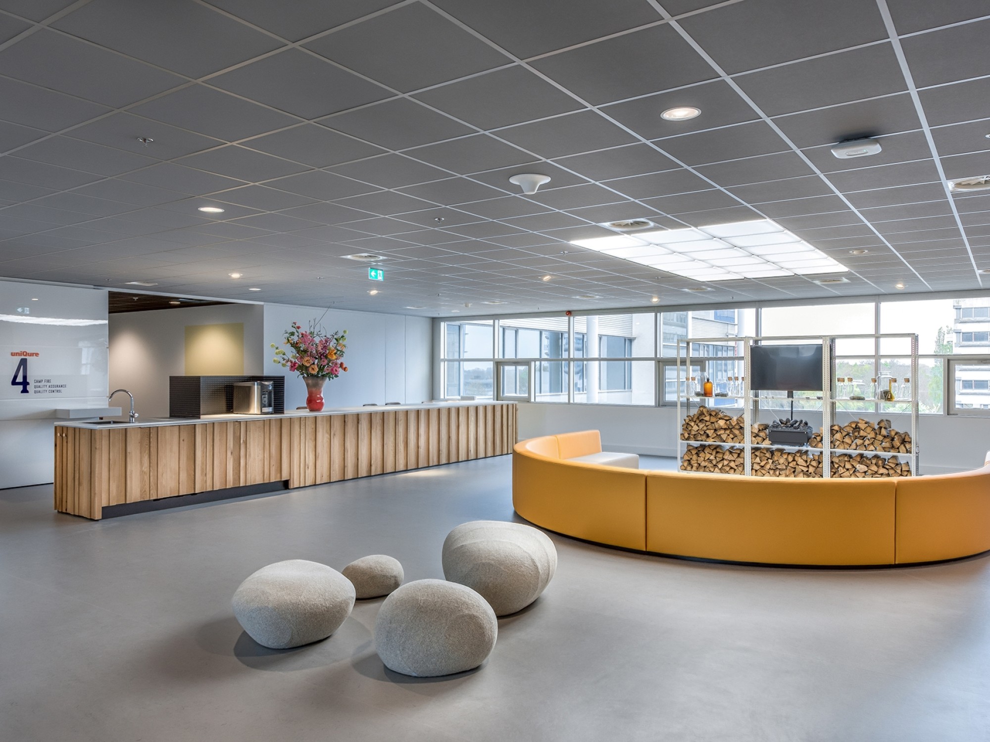 Een moderne ontvangsthal met een lange houten receptiebalie aan de linkerzijde. In het midden staat een ronde gele zitbank met een glazen vitrine erop. Op de voorgrond liggen enkele ronde zitpoefjes. Grote ramen zorgen voor veel natuurlijk licht in de rui