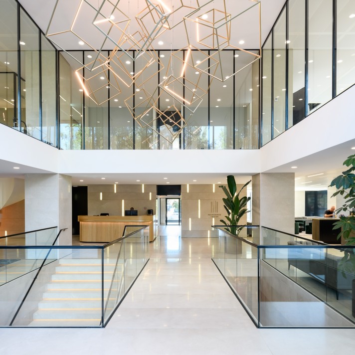 A bright, modern office lobby with a central staircase and glass railings. A large geometric light installation hangs from the ceiling above the open atrium. A reception desk and tall indoor plants are visible on the ground floor, surrounded by glass-wall