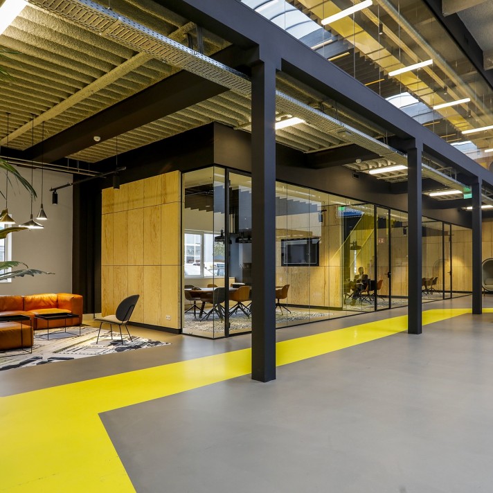 A modern industrial-style office space with black steel columns and exposed ceiling structures. A glass meeting room with wooden walls is centered in the space, while an orange lounge seating area is visible on the left. A bright yellow strip runs along t