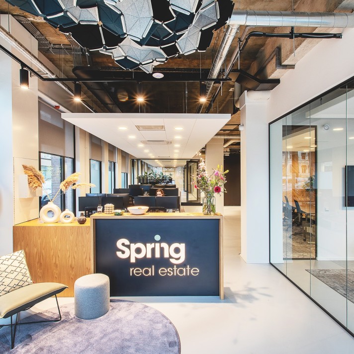 Een moderne kantoorentree met een receptiebalie waarop het logo “Spring real estate” staat. Boven de balie hangt een opvallende designlamp aan een industrieel plafond met zichtbare leidingen. Rechts is een glazen vergaderruimte zichtbaar en de ruimte heef