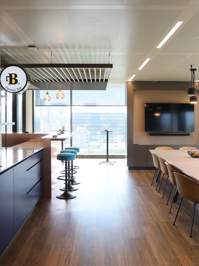 Moderne kantoorkeuken met donkerblauwe kasten, een kookeiland met barkrukken, een lange eettafel met stoelen, hanglampen en grote ramen met uitzicht.