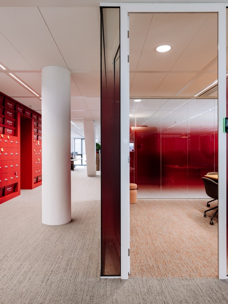 Moderne kantoorruimte met rode kastenwand, glazen vergaderruimte met rode wanden en een witte kolom in de gang.